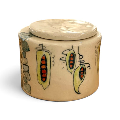 A handmade ceramic lidded container with a beige stoneware finish, decorated with hand-painted motifs of yellow-green peaches with red seeds and gray stone-like shapes on the side, set against a plain white background.