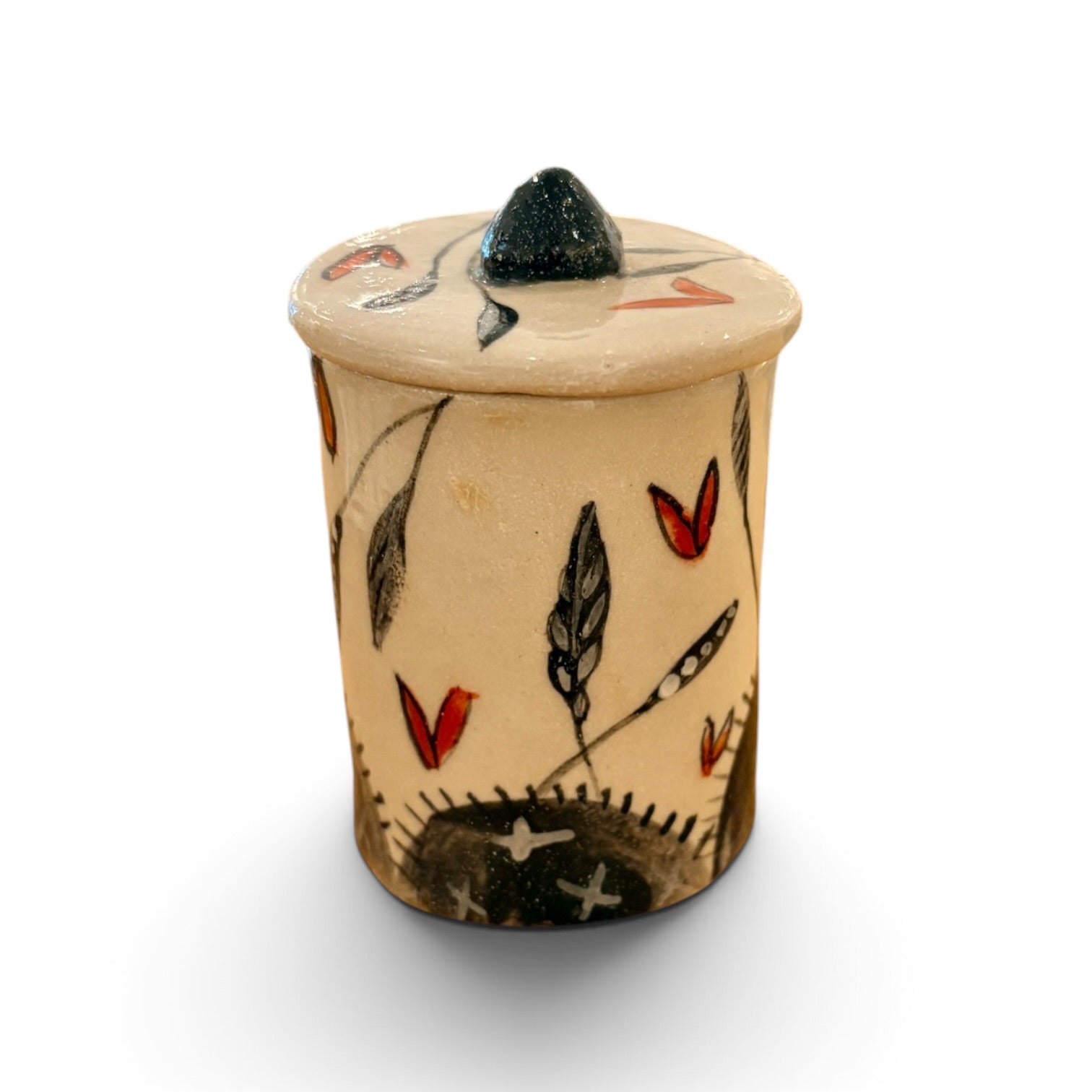 A small handmade ceramic lidded container with a beige body, decorated with hand-painted black and red motifs, and topped with a dark green pointed lid, shown on a white background.