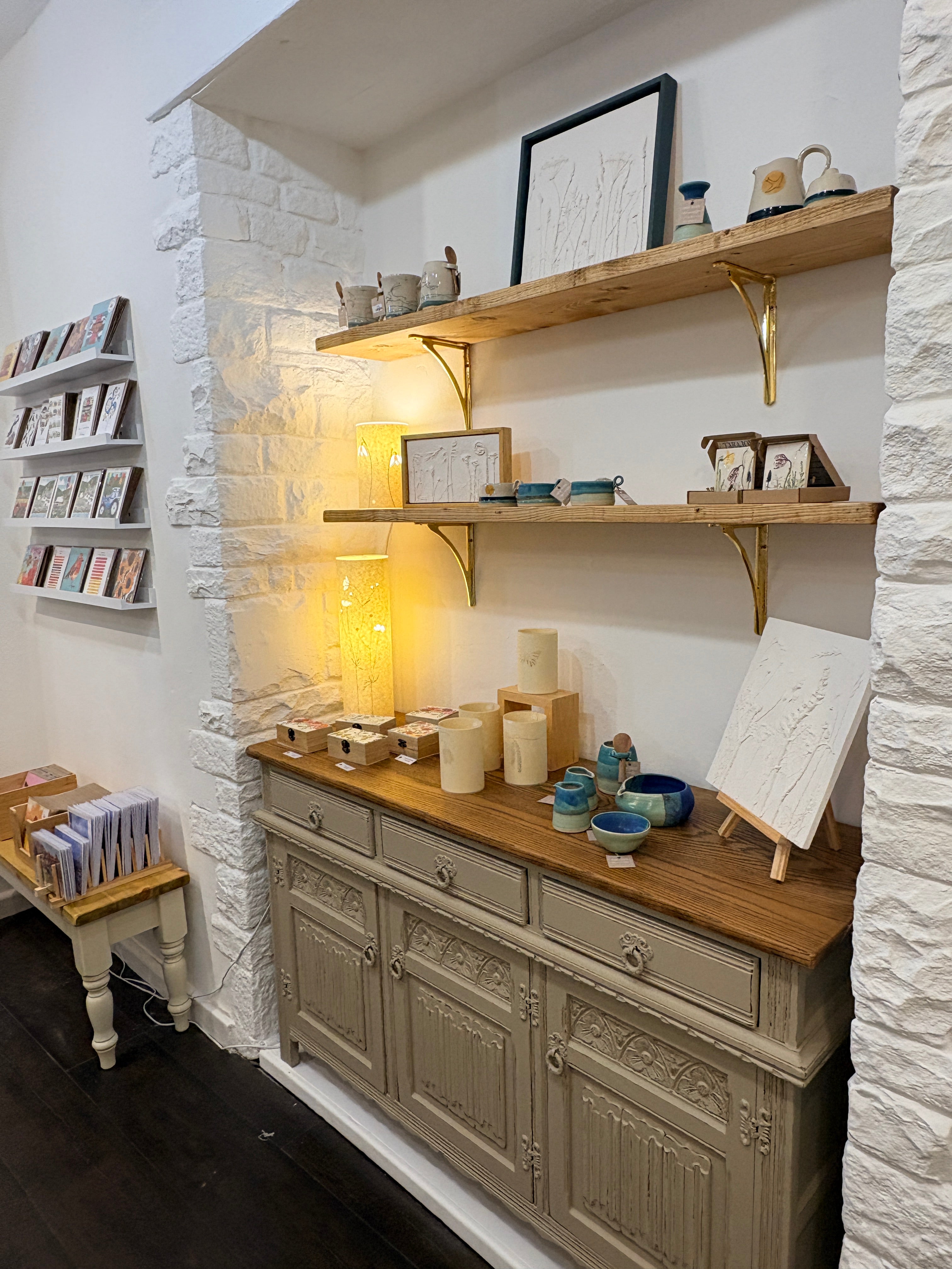 A styled interior scene showing a rustic wood mantel shelf with gold brackets, warm string lights, candles, framed art, and a carved wooden cabinet against a white brick fireplace.