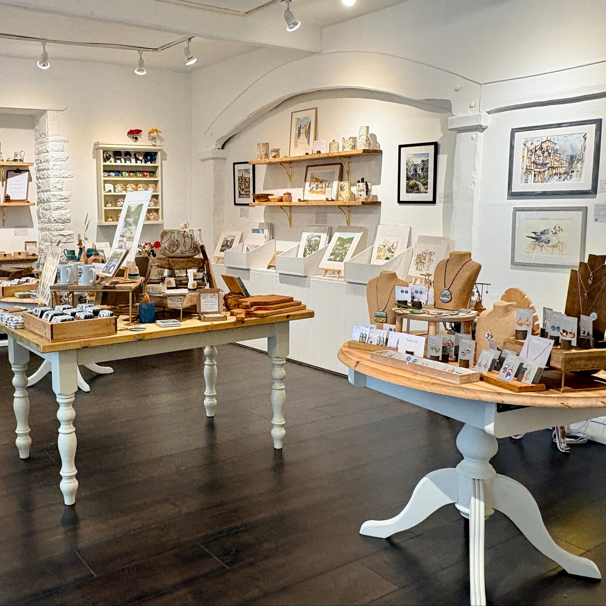 A rustic gift shop interior with dark wood floors, white walls, and display tables showcasing handmade wooden items, framed art, and small decorative products.
