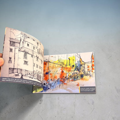 An open art book or sketchbook lying on a light gray surface, showing two illustrated pages: one black-and-white architectural drawing and one colorful street scene with orange, yellow, blue, and white watercolor tones.