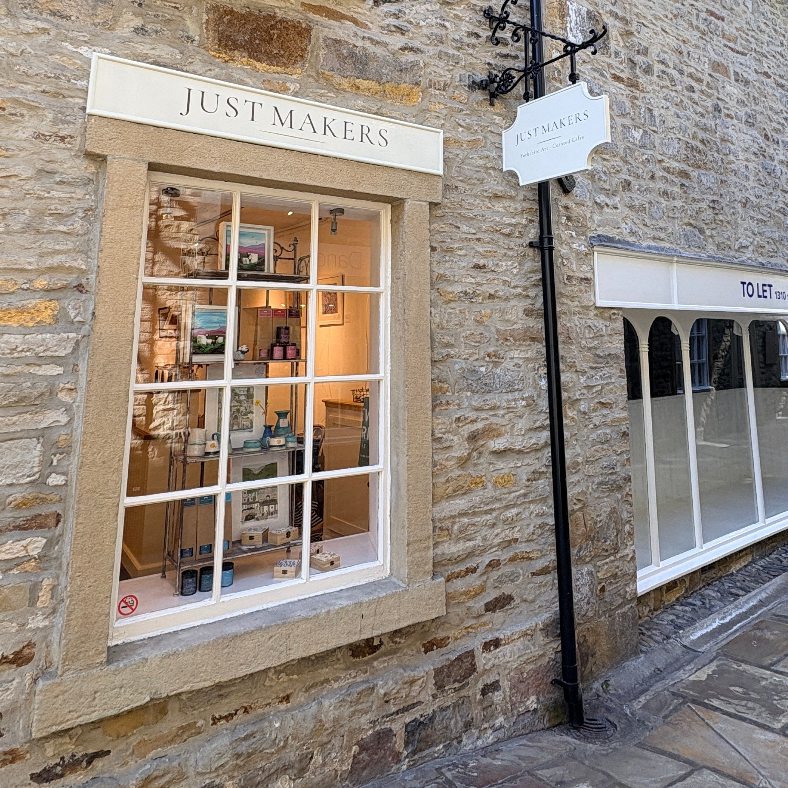 Just Makers boutique storefront on a stone wall, with a large white-framed window displaying shelves and products inside, a black hanging sign, and a neighboring shop window partially visible.