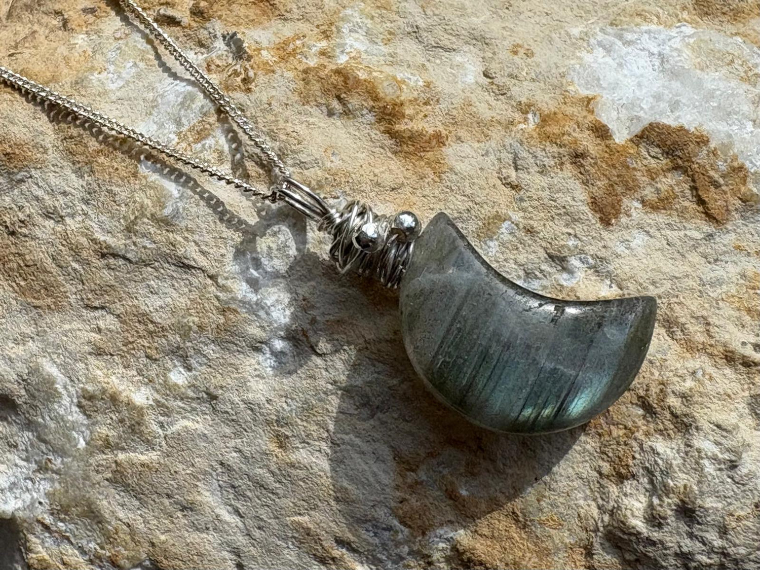 A silver necklace with a polished crescent moon pendant made of iridescent labradorite, resting on a textured natural stone surface.