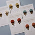 Six pairs of Collette Haigh handmade earrings displayed on their white backing cards, featuring various earthy tones including terracotta, dusty rose, sage green, cream, and light tan, with bronze leaf-shaped metal accents.