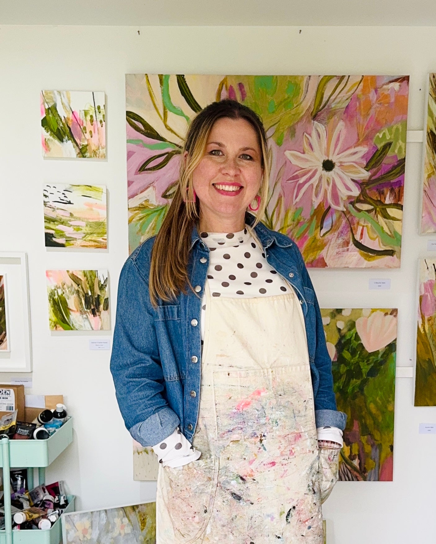 A smiling woman with blonde hair stands in an art studio wearing a blue denim jacket over a white top with black polka dots and a paint-splattered cream apron. She is standing in front of a large colorful floral painting on a white wall, with smaller paintings hanging to her left.