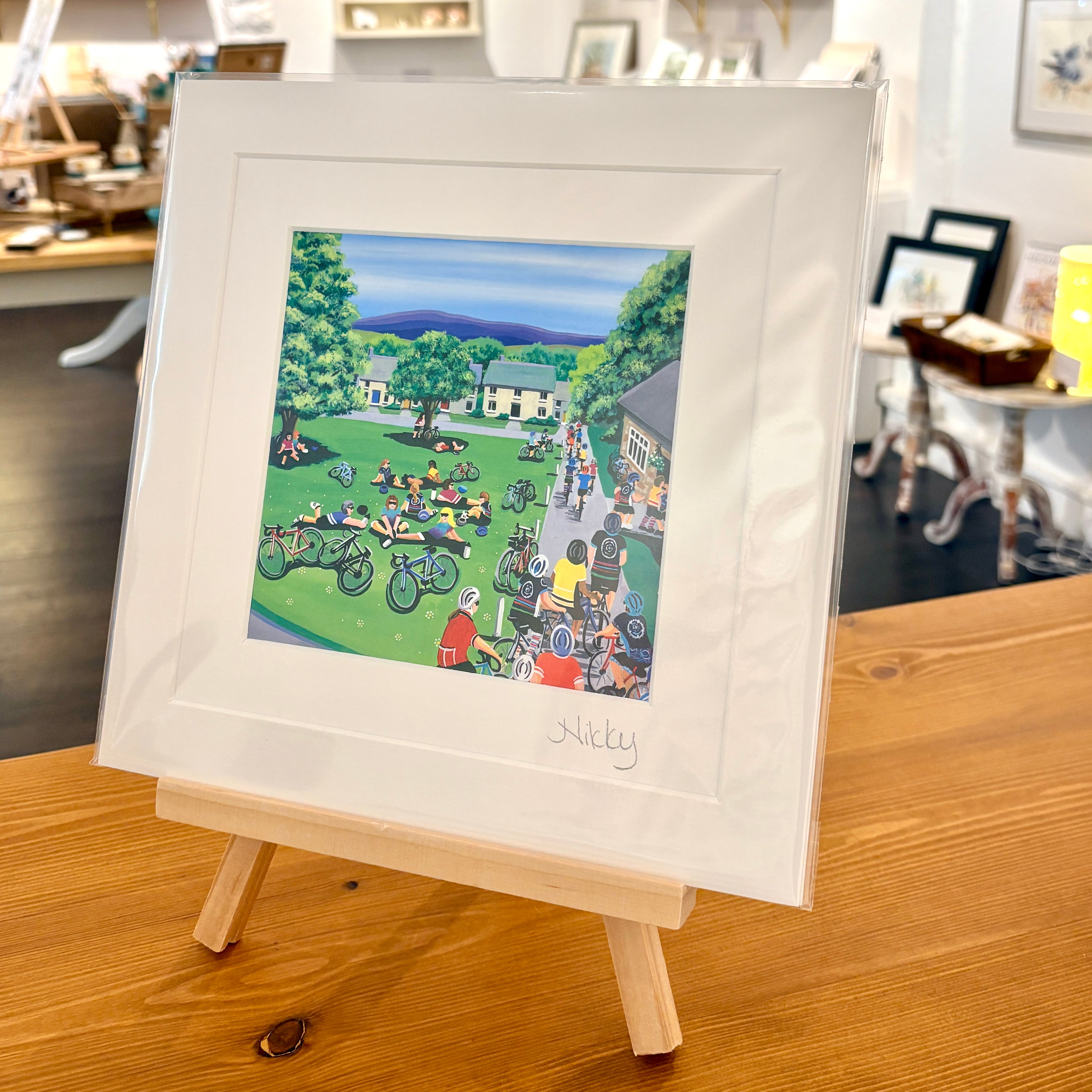 A framed art print featuring a colorful illustration of a village scene with people, bicycles, and green lawns, displayed on a small wooden easel in a shop setting.