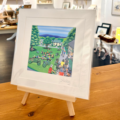 A framed art print featuring a colorful illustration of a village scene with people, bicycles, and green lawns, displayed on a small wooden easel in a shop setting.