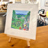A framed art print featuring a colorful illustration of a village scene with people, bicycles, and green lawns, displayed on a small wooden easel in a shop setting.