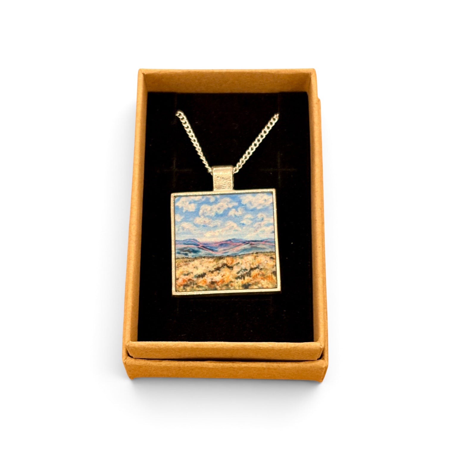A square pendant necklace displayed inside an open tan gift box with a black velvet insert. The pendant has a gold-tone chain and border, with a colorful landscape painting in the center showing blue sky, white clouds, and a golden field.