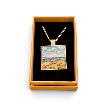 A square pendant necklace displayed inside an open tan gift box with a black velvet insert. The pendant has a gold-tone chain and border, with a colorful landscape painting in the center showing blue sky, white clouds, and a golden field.