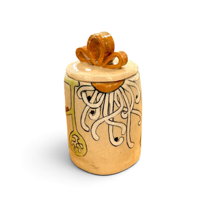 A handmade ceramic lidded jar with a beige body, hand-painted white tentacle-like pattern, yellow accents, and a glossy orange-brown lid ornament, shown on a white background.