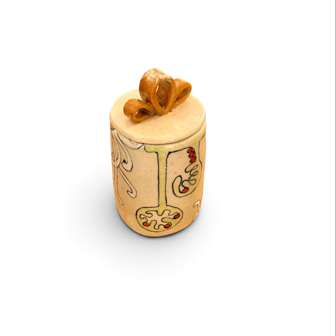 A small cylindrical decorative object with a lid, shown on a plain white background. The body is a warm beige/tan tone with painted ornamental motifs in muted green, red, and dark lines, topped with a folded golden-brown ribbon or bow on the lid.