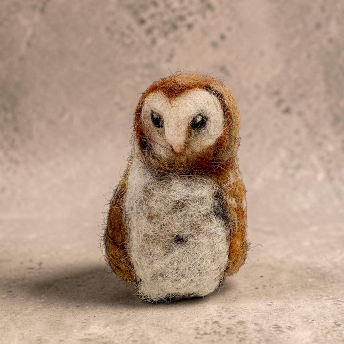 A small felted wool owl figurine with brown and white feathers, standing against a textured beige background.