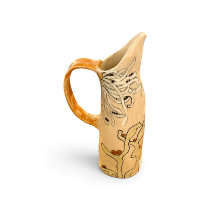 A handmade ceramic pitcher with a warm beige body, a glossy orange-brown handle and rim, and hand-painted botanical designs in black and brown on the side, set against a plain white background.