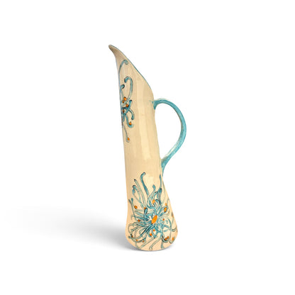 A tall, slender ceramic pitcher with a curved handle, featuring a cream-colored glaze decorated with turquoise blue floral and scroll patterns and small orange accents, set against a plain white background.