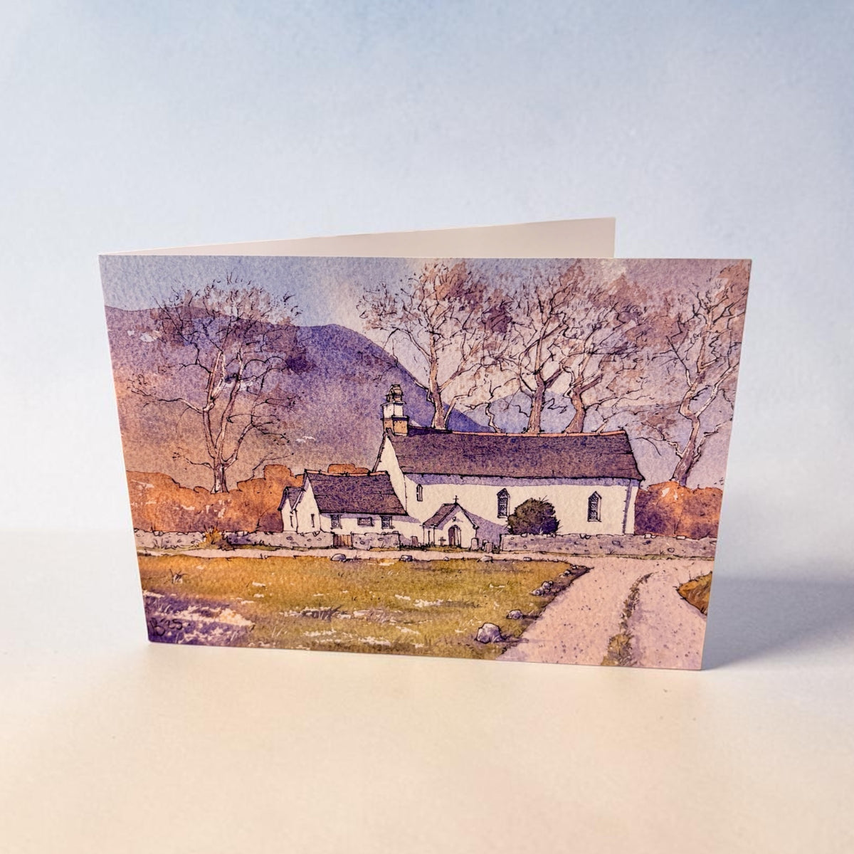 A square art print or card featuring a watercolor-style landscape of a white church with bare trees, a dirt path, and distant hills, photographed standing upright against a pale blue and beige studio background.
