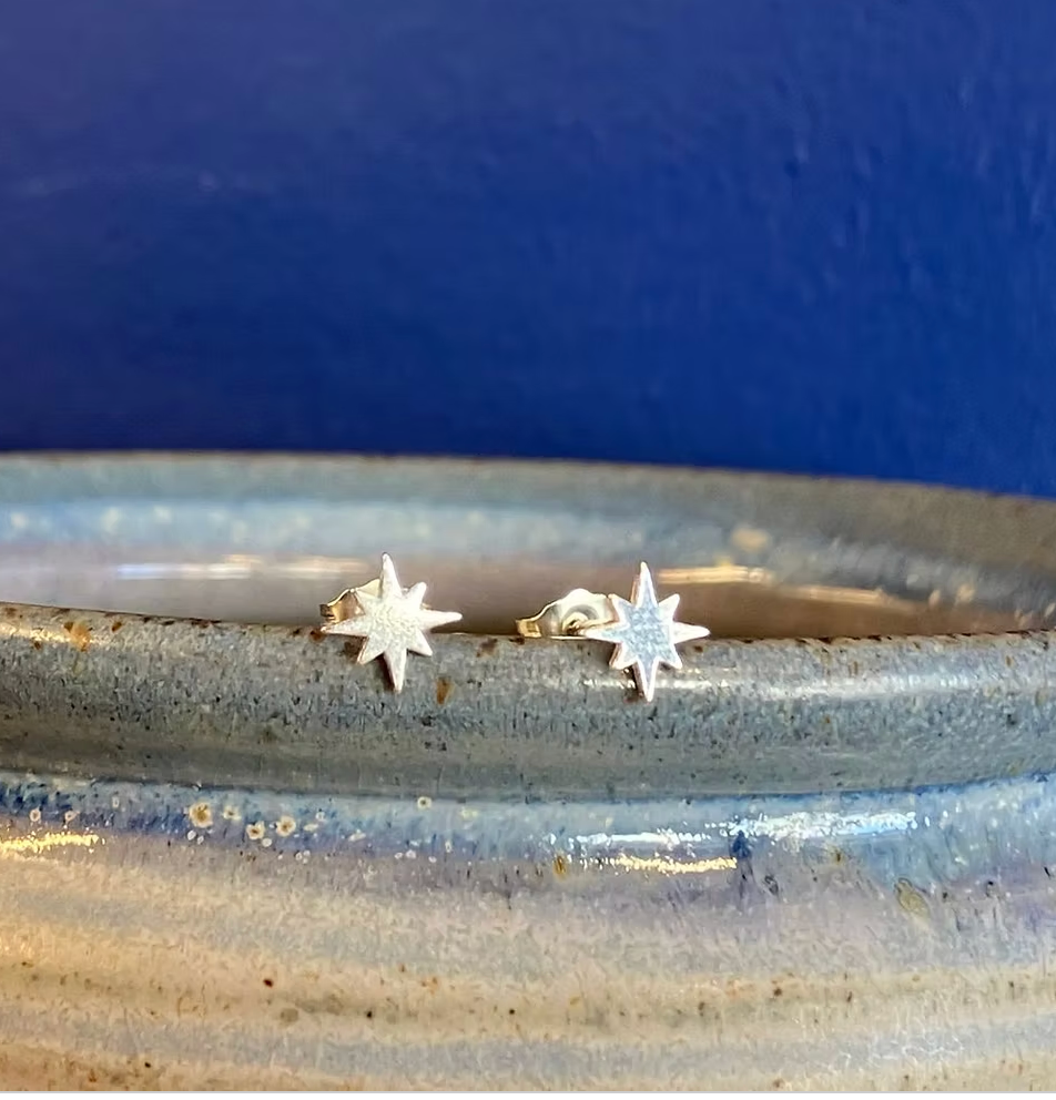 A pair of small silver starburst stud earrings resting on the rim of a speckled ceramic dish against a dark blue background.