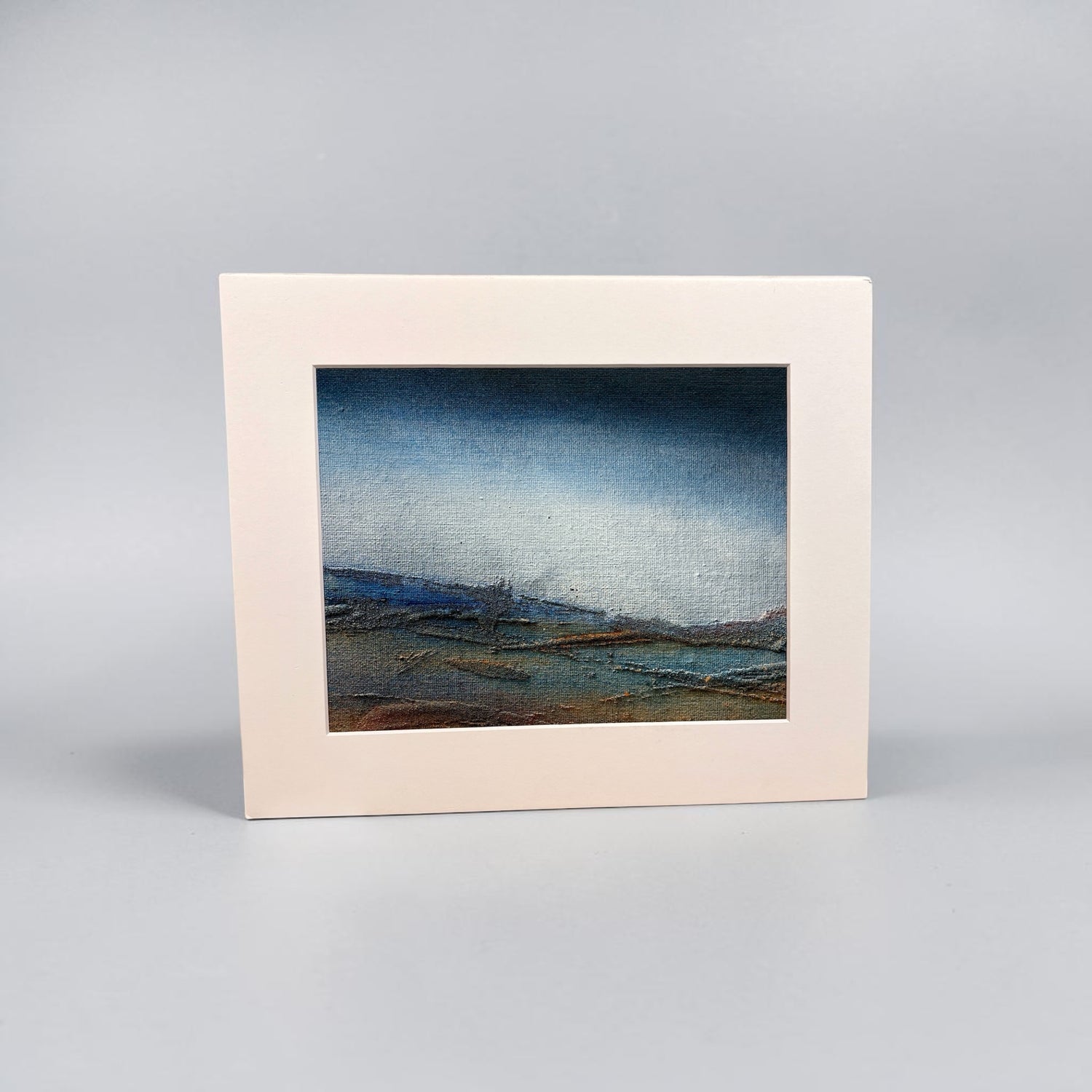 A framed landscape art print with a wide cream-colored border and a textured painting depicting a moody blue sky over a low, dark coastal scene, set against a neutral gray background.