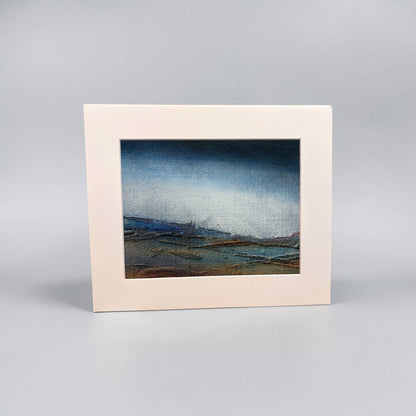 A framed landscape art print with a wide cream-colored border and a textured painting depicting a moody blue sky over a low, dark coastal scene, set against a neutral gray background.