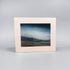 A framed landscape art print with a wide cream-colored border and a textured painting depicting a moody blue sky over a low, dark coastal scene, set against a neutral gray background.