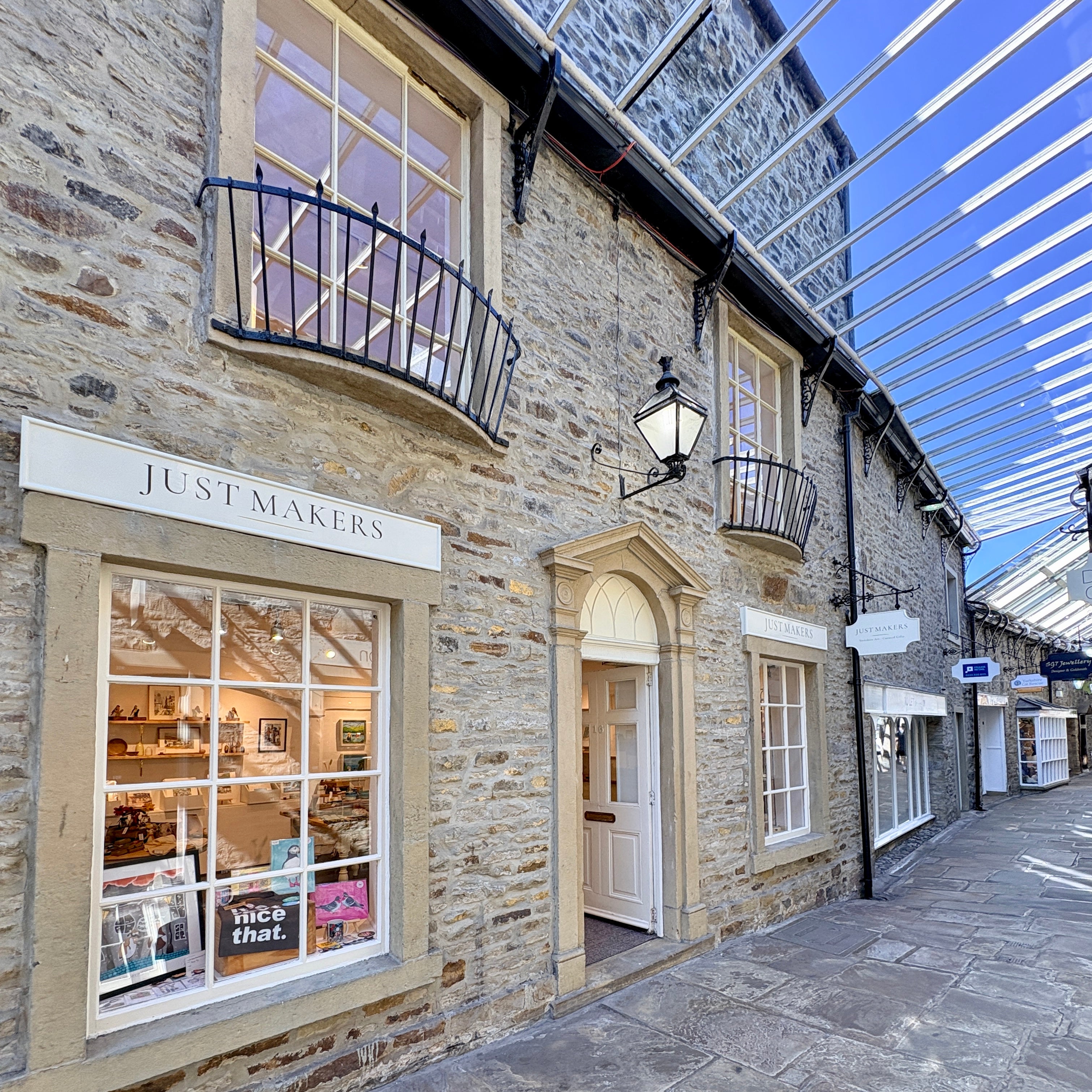A stone shopfront building with a sign reading JUST MAKERS above a large window display, set in a covered outdoor alley with a glass roof and stone pavement.