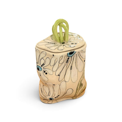 A handcrafted ceramic canister with a lid, featuring a beige base decorated with white floral patterns outlined in black, accented with small blue and black dots, and topped with a light green loop handle, shown on a white background.