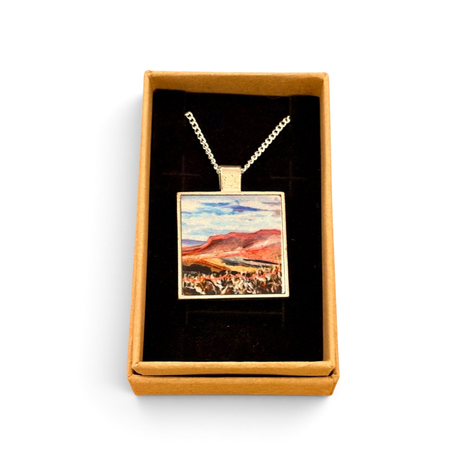 A square pendant necklace displayed in an open gift box. The pendant has a silver-toned border and chain, with a colorful landscape painting inside featuring blue sky, red hills, and a white foreground.