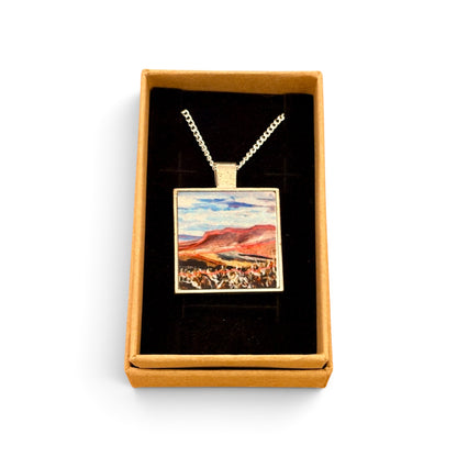 A square pendant necklace displayed in an open gift box. The pendant has a silver-toned border and chain, with a colorful landscape painting inside featuring blue sky, red hills, and a white foreground.