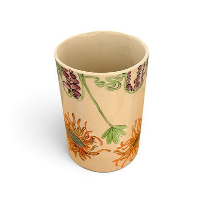 A beige ceramic cup decorated with orange chrysanthemums and green leaves, with purple and green floral accents near the rim, shown on a white background.