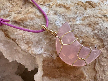 A pink crescent moon pendant necklace made from polished rose quartz wrapped in gold wire, hanging from a purple cord against a textured beige rock background.