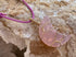 A pink crescent moon pendant necklace made from polished rose quartz wrapped in gold wire, hanging from a purple cord against a textured beige rock background.