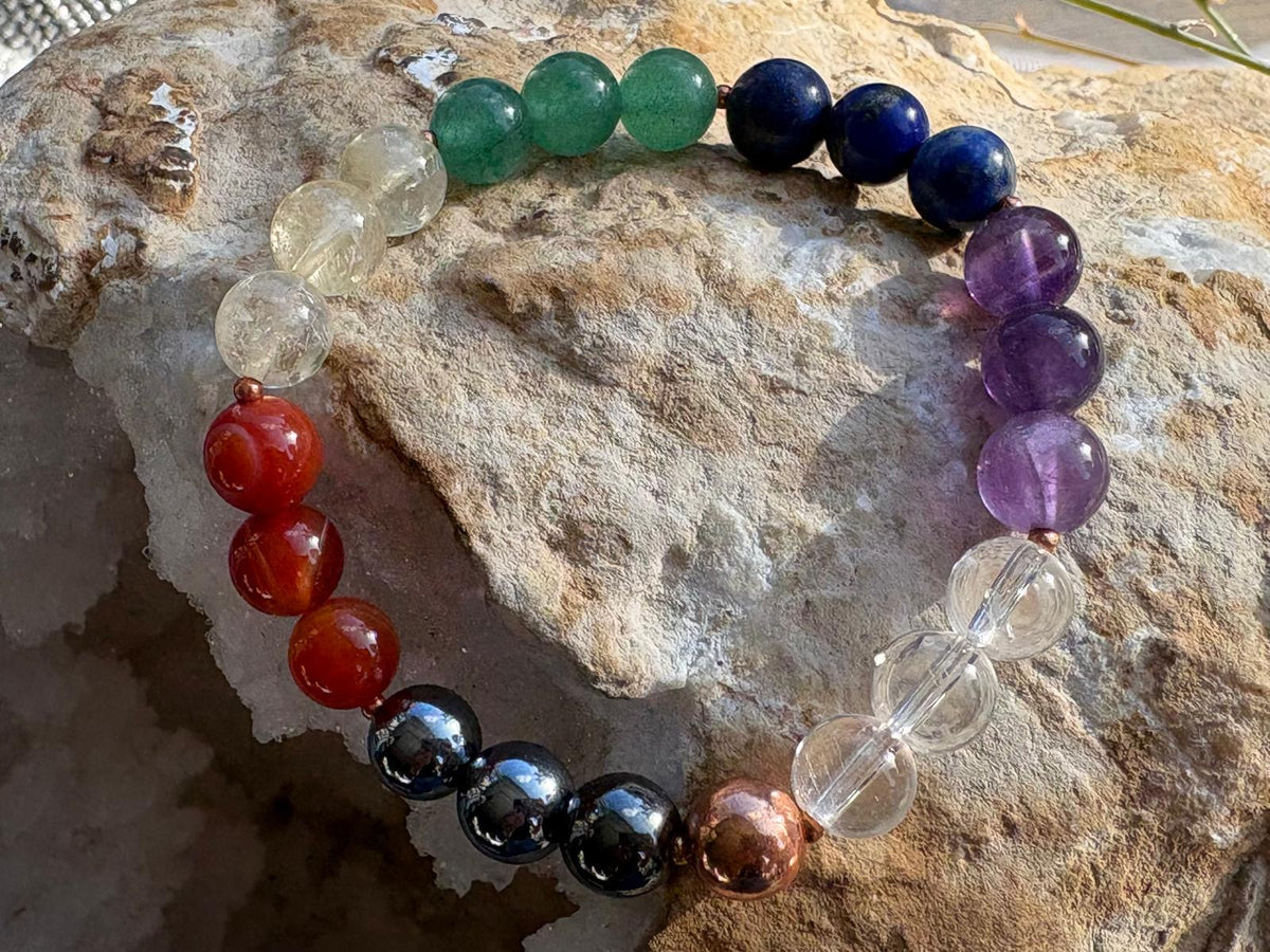A multicolor chakra bead stretch bracelet arranged in a circle on a textured beige rock. The beads represent the seven chakras in color order: red, orange, dark blue, black, copper, clear, purple, white, and green.