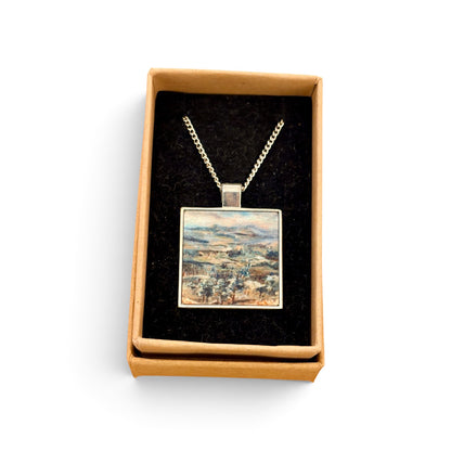 A square pendant necklace displayed in an open tan gift box with a black velvet insert. The pendant features a colorful landscape painting with blue, green, and beige tones, suspended from a silver-toned chain.