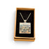 A square pendant necklace displayed in an open tan gift box with a black velvet insert. The pendant features a colorful landscape painting with blue, green, and beige tones, suspended from a silver-toned chain.