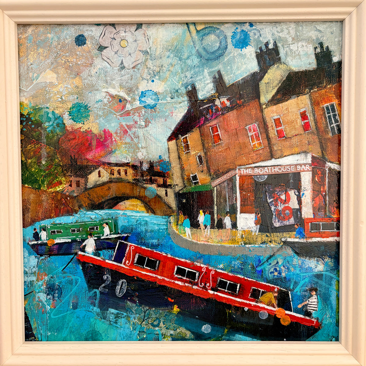 An expressive, colorful painting of a canal-side town scene with narrowboats on blue water, brick buildings, a bridge, and a pub, shown in a light wood frame.