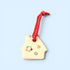 A small house-shaped ceramic ornament in a warm cream color, decorated with colorful painted flowers and a red heart, hanging from a red ribbon with white dots, set against a solid light blue background.