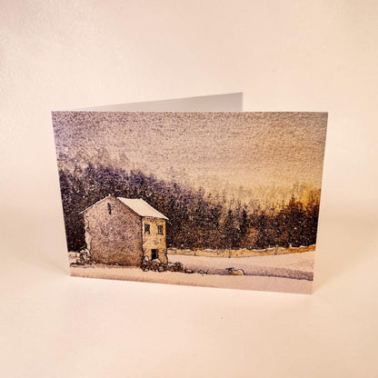 A folded greeting card standing upright on a light beige surface, printed with a winter landscape scene featuring a snow-covered stone house, dark trees, and a pale sky.