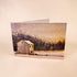 A folded greeting card standing upright on a light beige surface, printed with a winter landscape scene featuring a snow-covered stone house, dark trees, and a pale sky.