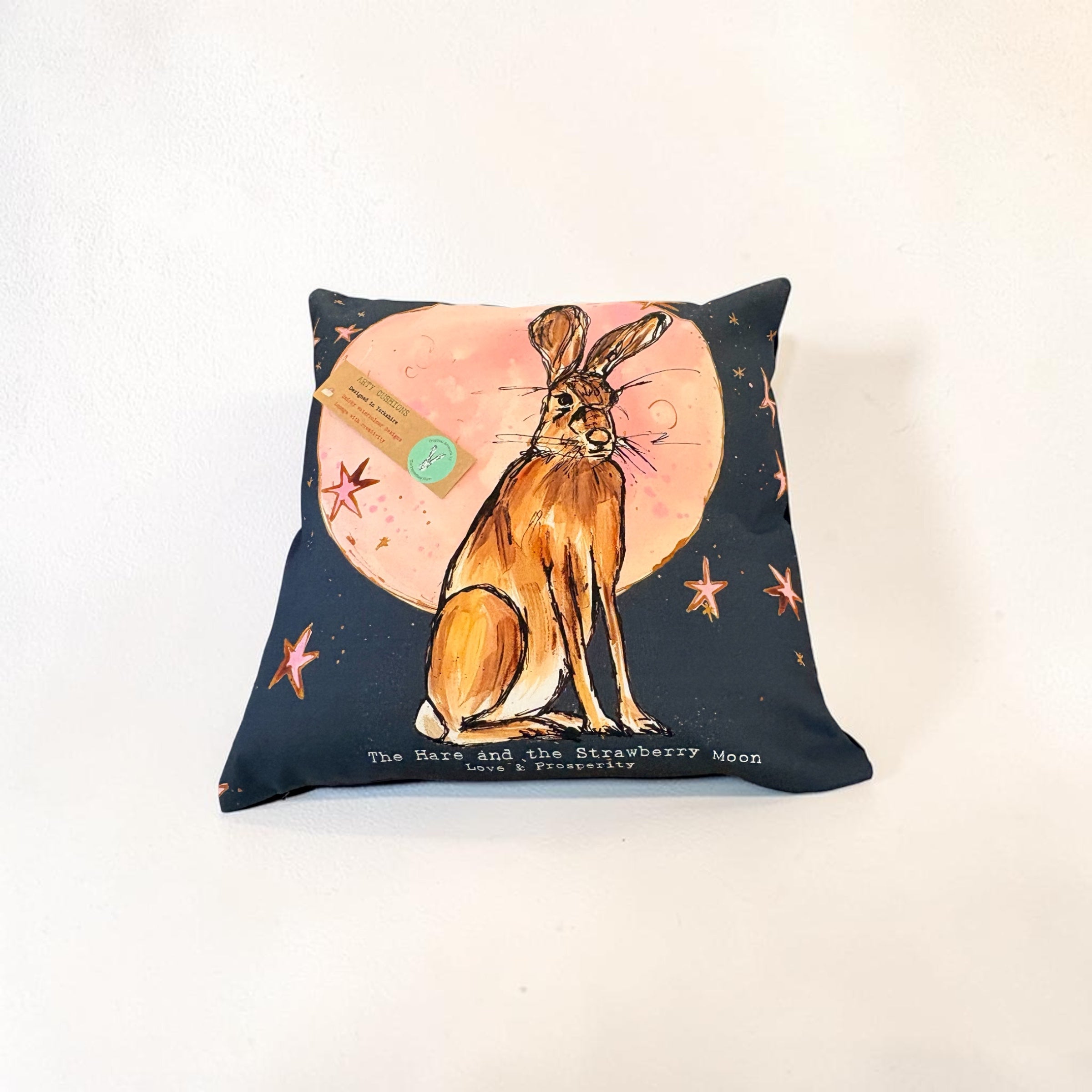 A square decorative pillow featuring a watercolor-style illustration of a brown hare sitting in front of a pink moon, with small orange stars on a dark navy background. A small paper tag is attached to the pillow, and the text &