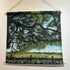 A fabric wall hanging scroll depicting a scenic landscape with a large tree, green field, flowers, and a swing, suspended by twine from wooden dowels on a white wall.