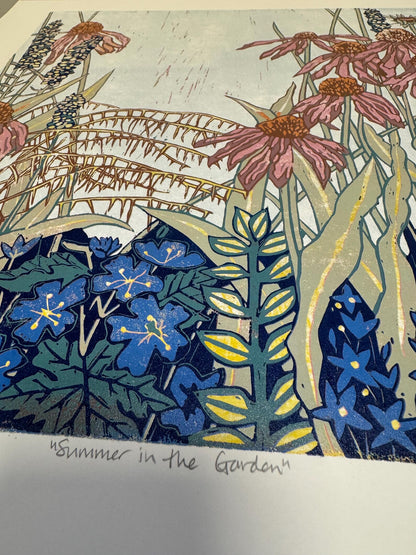 A close-up of a printed artwork or poster showing a garden scene with pink flowers, blue blossoms, green and yellow leaves, and a pale sky. Handwritten text at the bottom reads “Summer in the Garden.