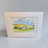 A framed watercolor landscape print featuring rolling green hills, a small house, trees, and a blue sky, displayed against a neutral light gray background.
