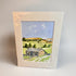 A framed watercolor landscape print showing a stone cottage on a grassy hillside under a blue sky, photographed against a neutral light gray background.