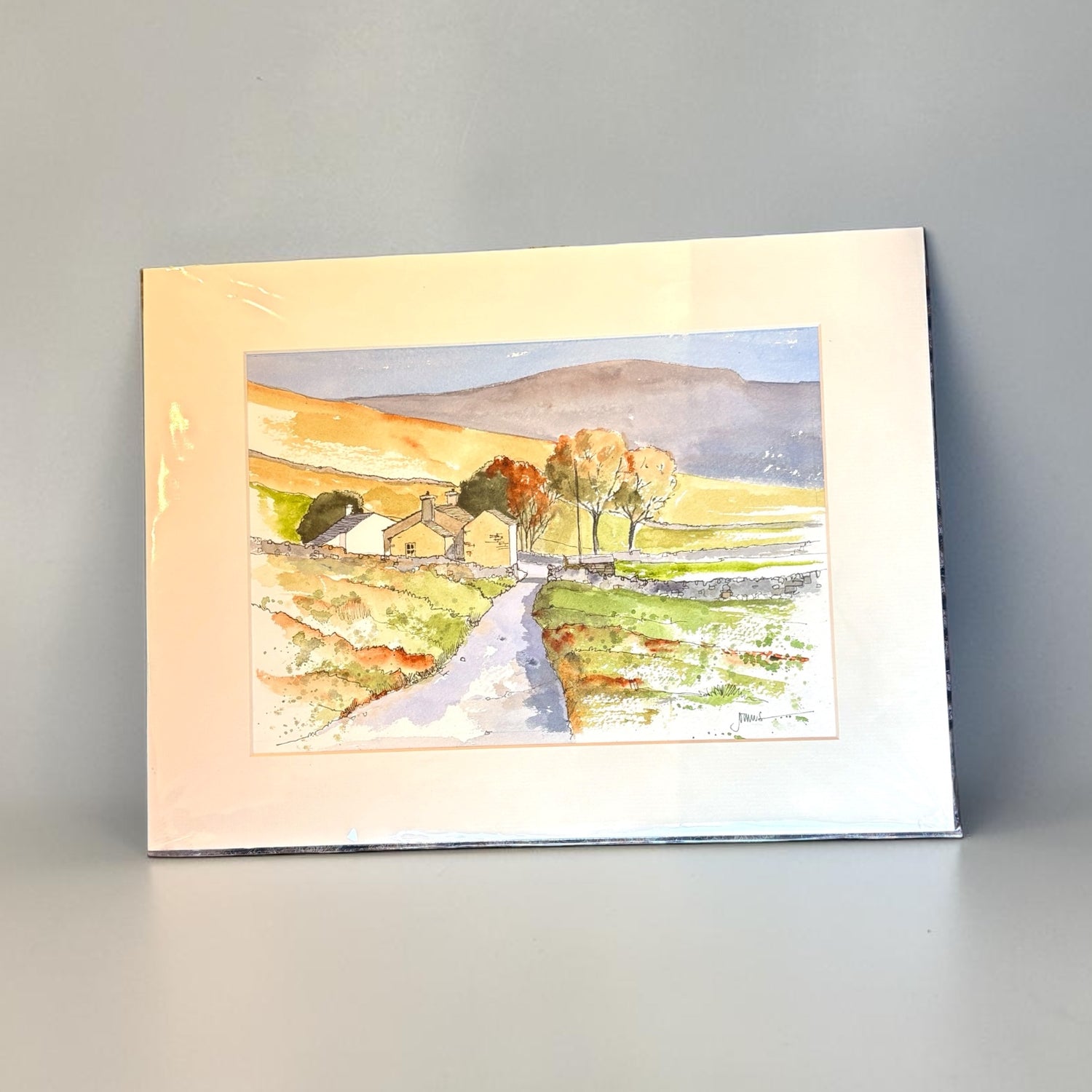 A watercolor landscape art print depicting a rural scene with a winding path, small cottages, trees, and rolling hills, shown with a wide cream mount against a neutral gray background.