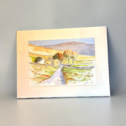 A watercolor landscape art print depicting a rural scene with a winding path, small cottages, trees, and rolling hills, shown with a wide cream mount against a neutral gray background.