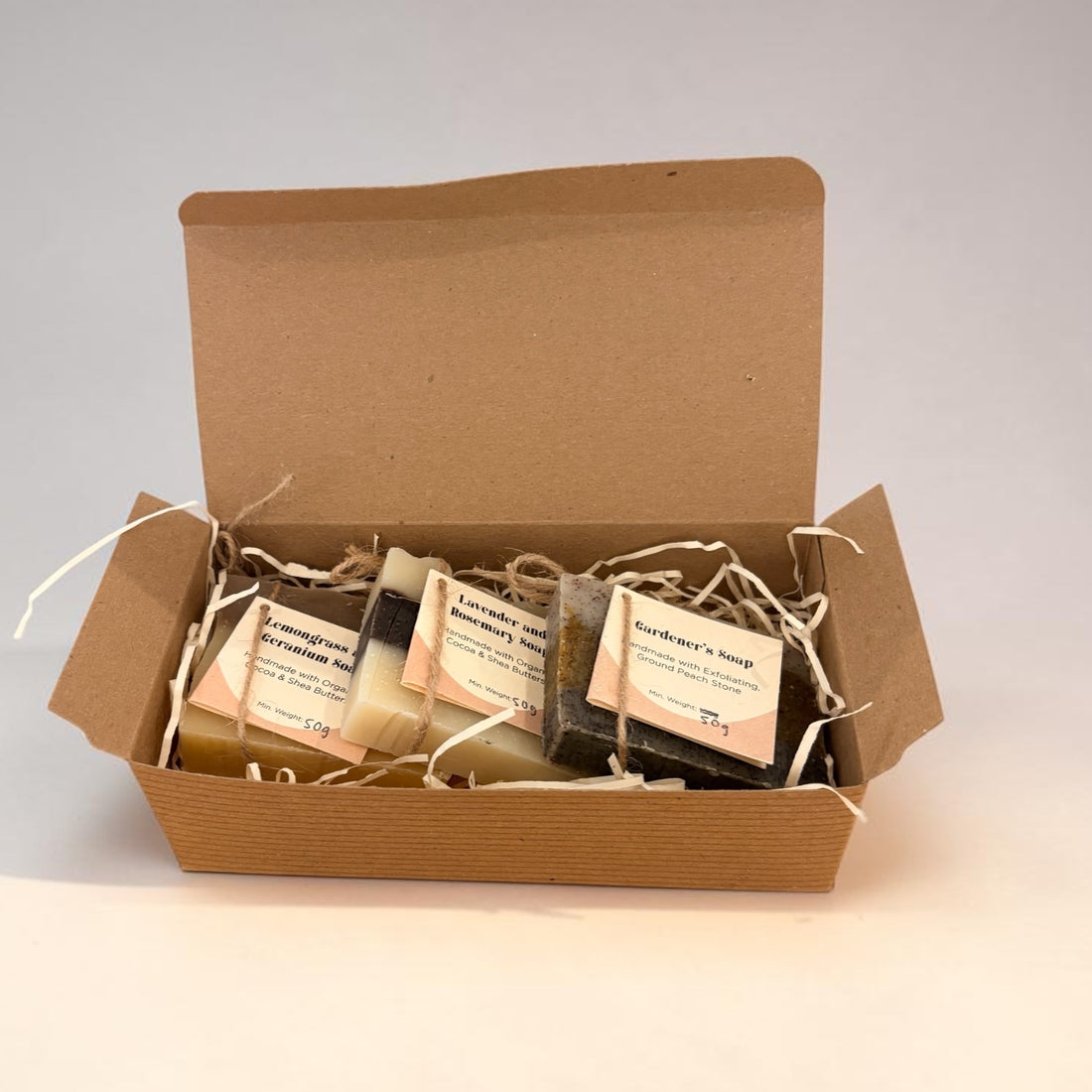 An open brown kraft gift box filled with several small wrapped soap bars on a plain light background. The soaps are individually wrapped in clear plastic with cream labels, and the box is lined with white paper shred.