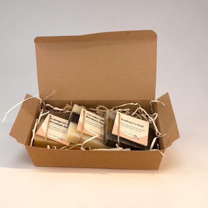 An open brown kraft gift box filled with several small wrapped soap bars on a plain light background. The soaps are individually wrapped in clear plastic with cream labels, and the box is lined with white paper shred.