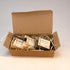 An open brown kraft gift box filled with several small wrapped soap bars on a plain light background. The soaps are individually wrapped in clear plastic with cream labels, and the box is lined with white paper shred.