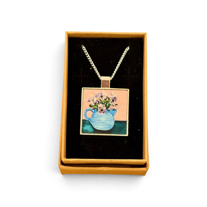 A square pendant necklace displayed in an open tan gift box with a black velvet insert. The pendant features a colorful illustration of a blue teapot filled with flowers against a peach and teal background, suspended from a silver-toned chain.