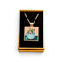 A square pendant necklace displayed in an open tan gift box with a black velvet insert. The pendant features a colorful illustration of a blue teapot filled with flowers against a peach and teal background, suspended from a silver-toned chain.